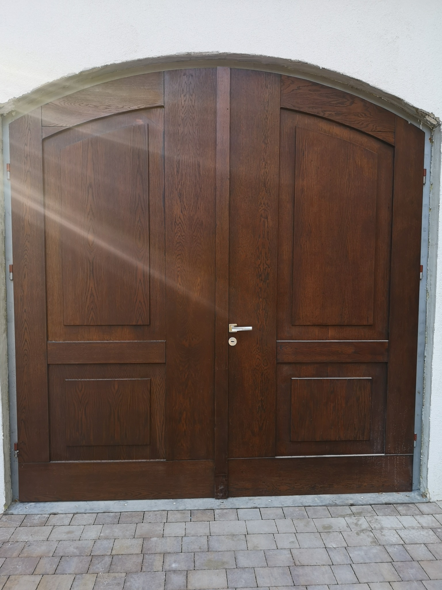 Dwuskrzydłowe, łukowe drzwi wejściowe z ciemnego drewna z metalową klamką, osadzone w białej ścianie, z widocznymi śladami prac montażowych wokół ościeżnicy.