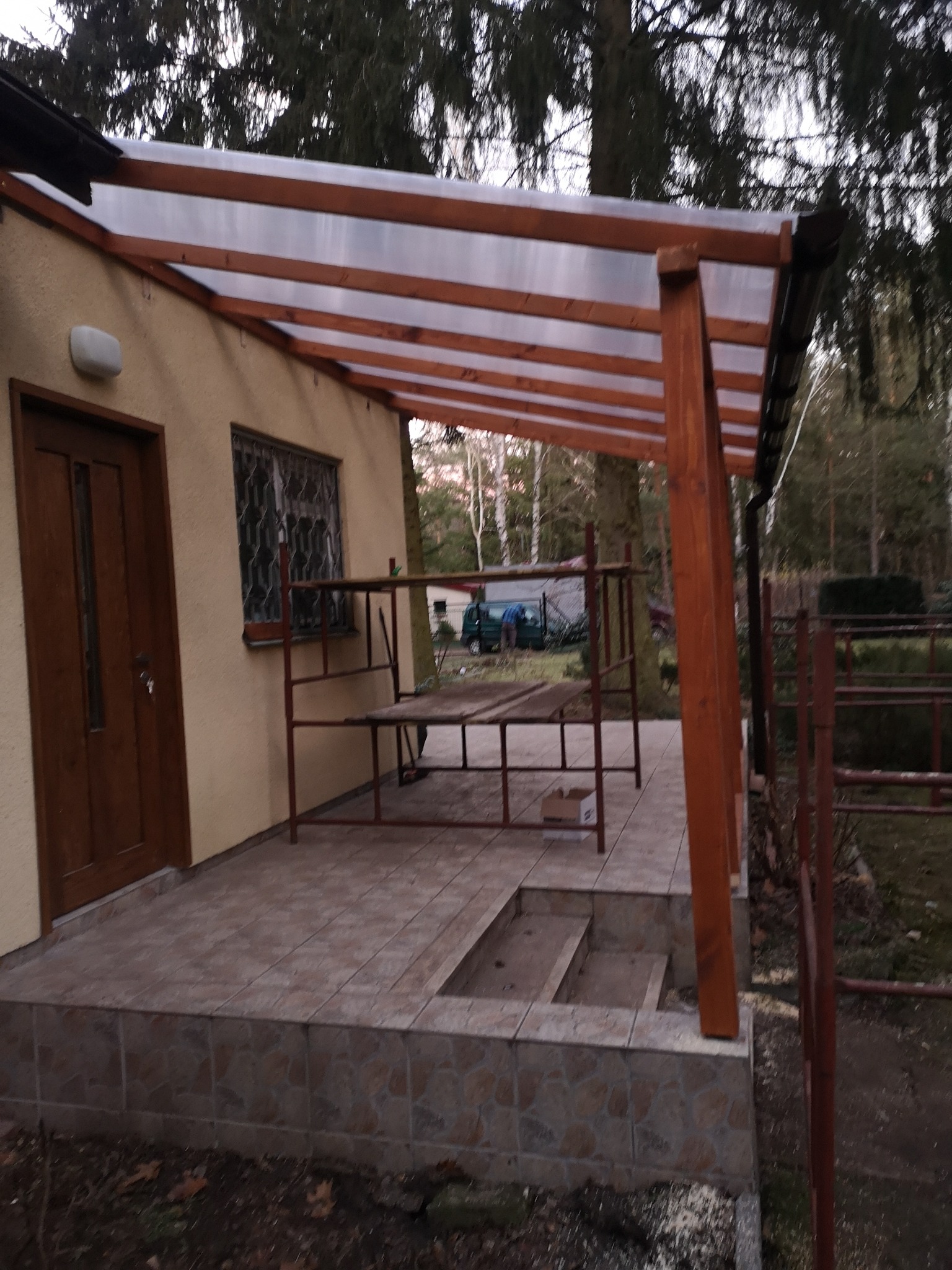 Zadaszenie tarasu z drewnianą konstrukcją i przezroczystym pokryciem, przylegające do budynku z brązowymi drzwiami i kratą w oknie, widoczne rusztowanie i drzewa w tle.