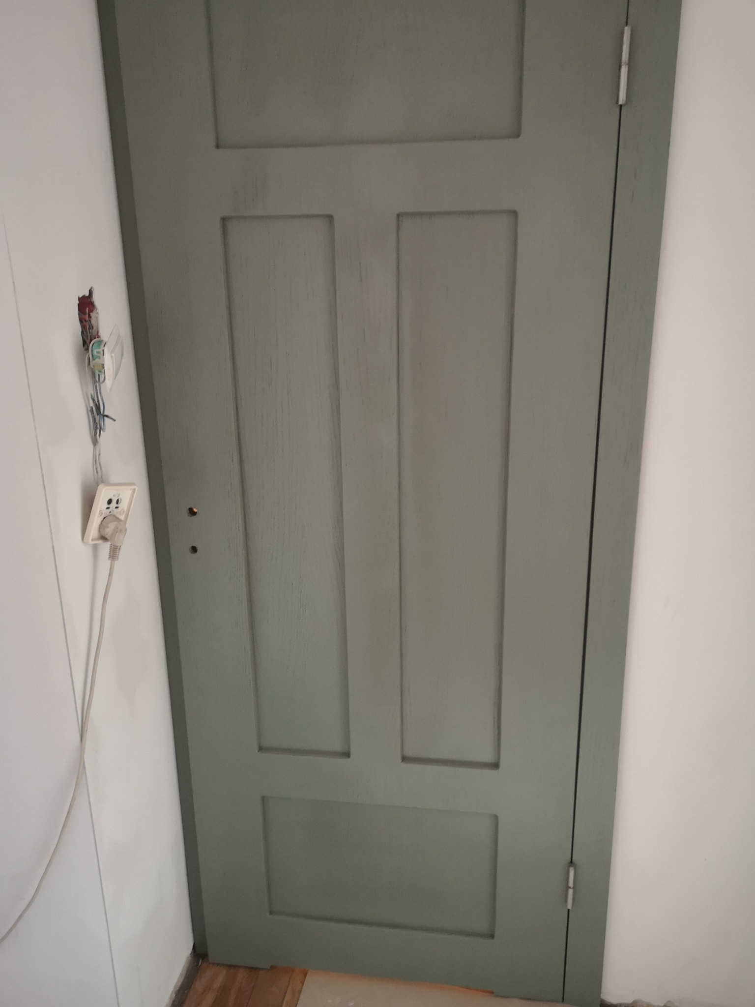Szare, wewnętrzne drzwi z trzema prostokątnymi panelami, widoczne zawiasy, obok ściana z wystającym gniazdkiem elektrycznym i przewodami.