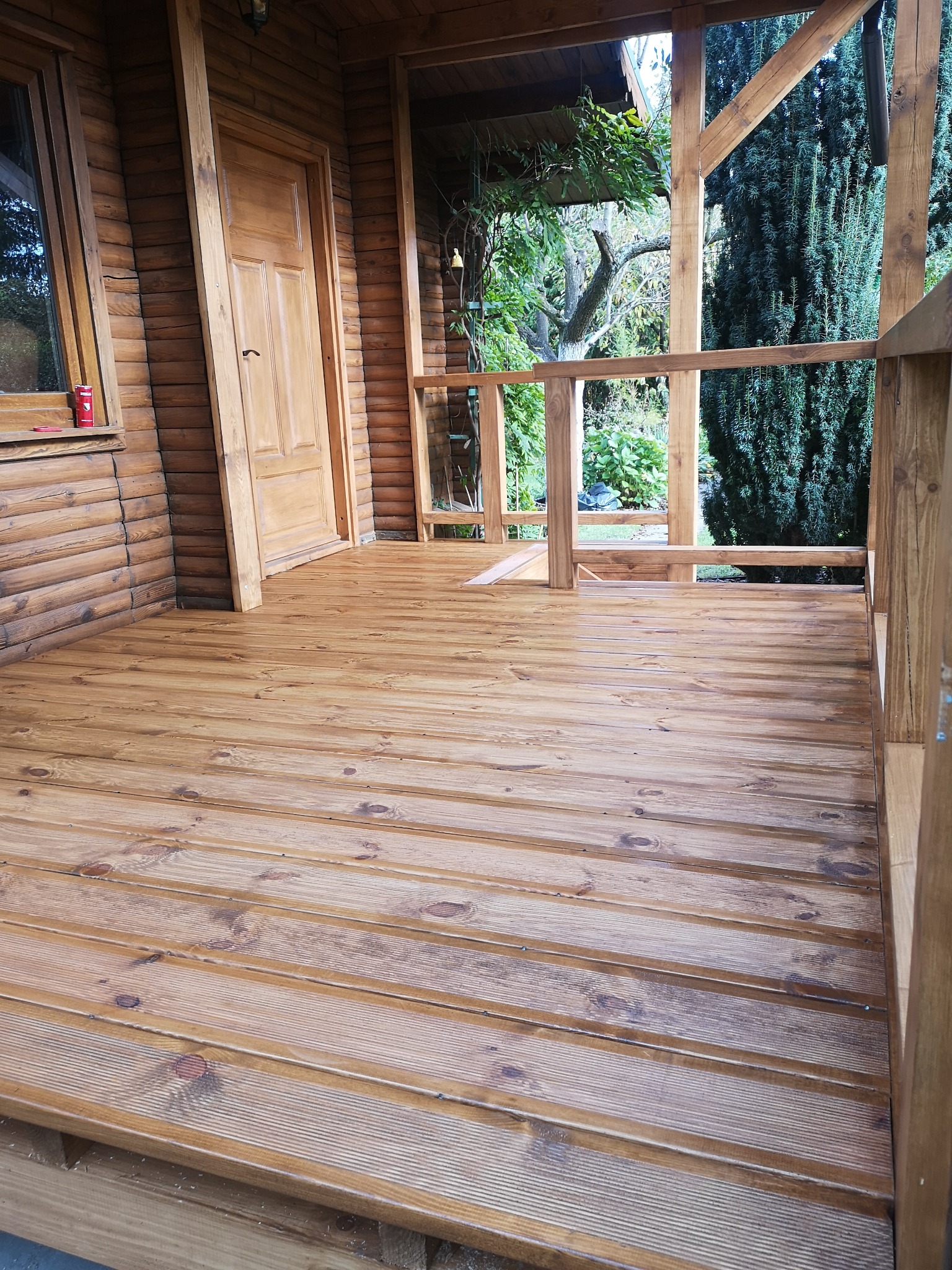 Drewniany taras z balustradą i widokiem na zieleń. Ściany z bali, drewniane drzwi. Naturalne oświetlenie podkreśla strukturę drewna.