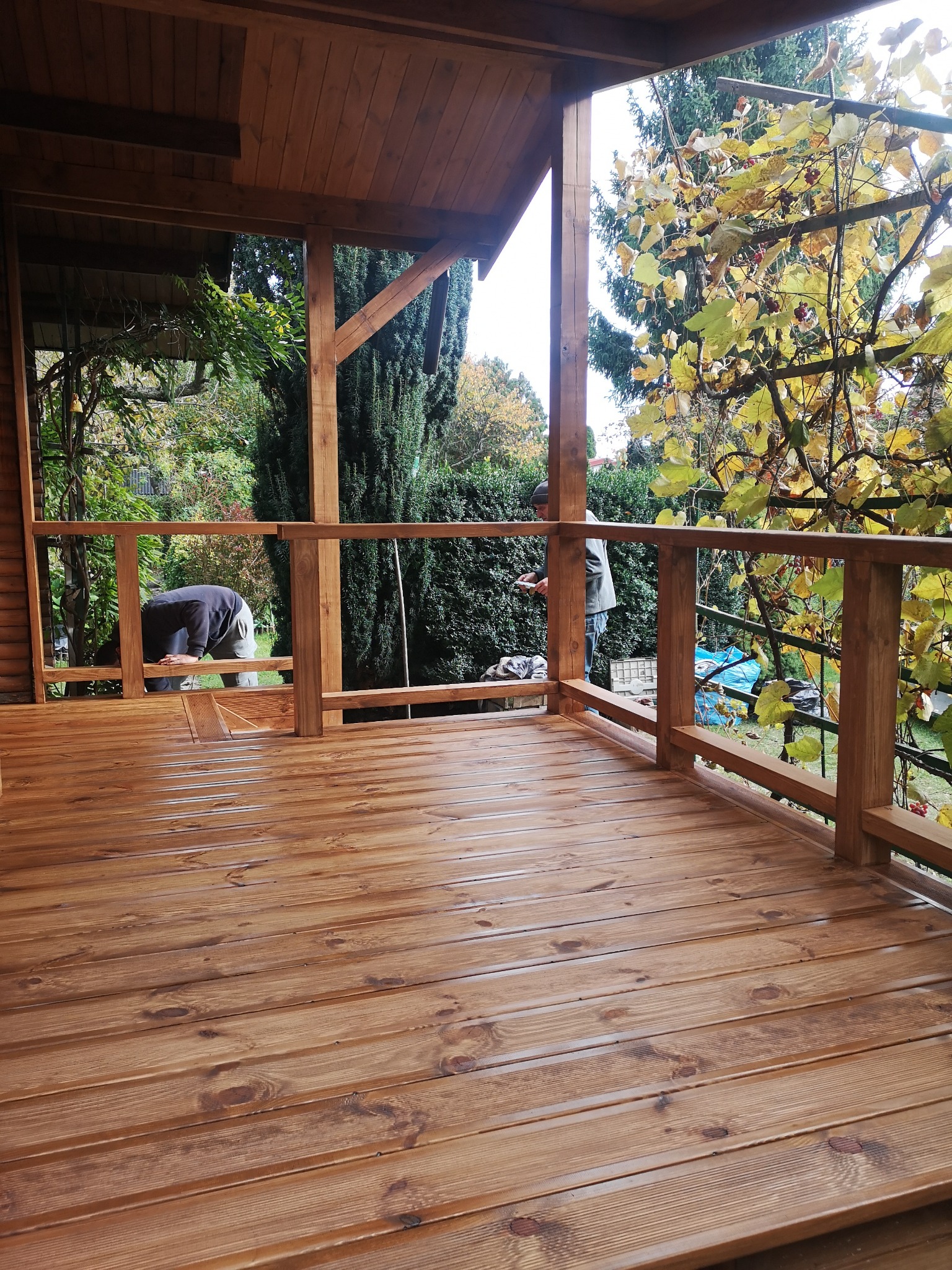 Drewniana konstrukcja z balustradą i podłogą, prawdopodobnie altana lub zadaszony taras, w trakcie prac wykończeniowych. Widoczni pracownicy. Tło: zieleń drzew.