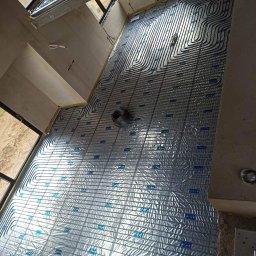 Hydroekipa. Specjaliści od mokrej roboty. - Rozłożona instalacja ogrzewania podłogowego na folii izolacyjnej z siatką, widok z góry na surowy stan wykończenia wnętrza z oknami.