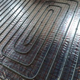 Hydroekipa. Specjaliści od mokrej roboty. - Szare rury ogrzewania podłogowego ułożone na folii z czerwoną siatką, zabezpieczone czarnymi klipsami, widok z góry.