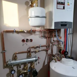 Nowoczesny piec gazowy Ariston ONE z rozbudowaną instalacją hydrauliczną z miedzianych rur, zbiornikiem wyrównawczym i pompą obiegową Grundfos, zamontowany na ścianie w jasnym pomieszczeniu.