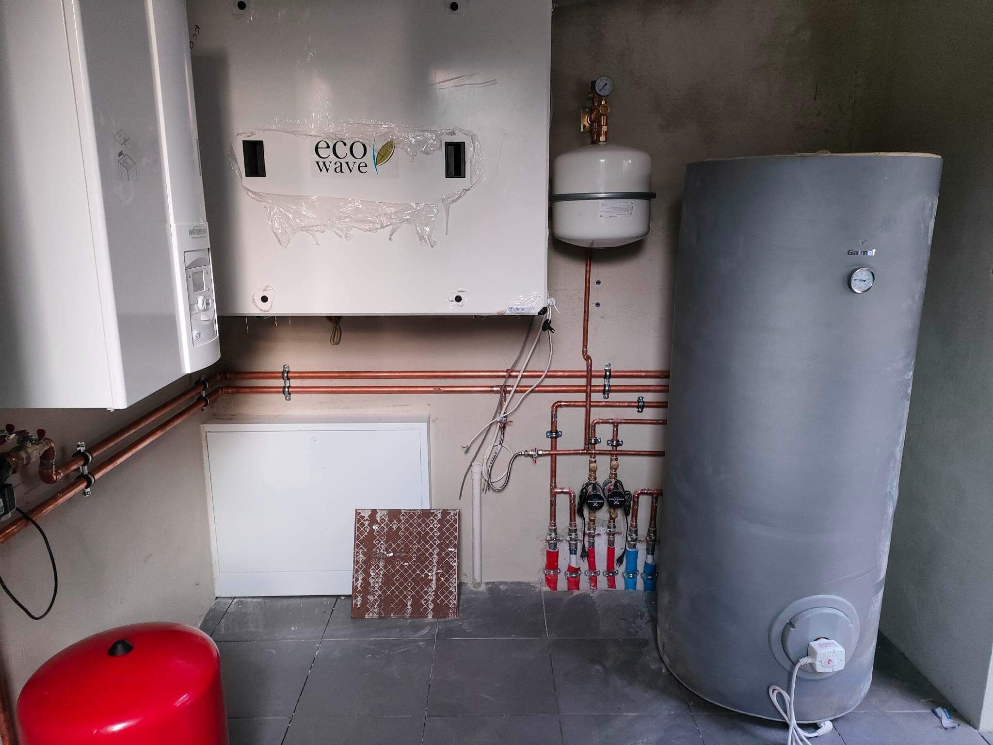 Nowa instalacja grzewcza z piecem Eco Wave, zbiornikiem akumulacyjnym i rurami miedzianymi, widoczne zawory i manometry, na podłodze płytki. Piec jeszcze w folii ochronnej.