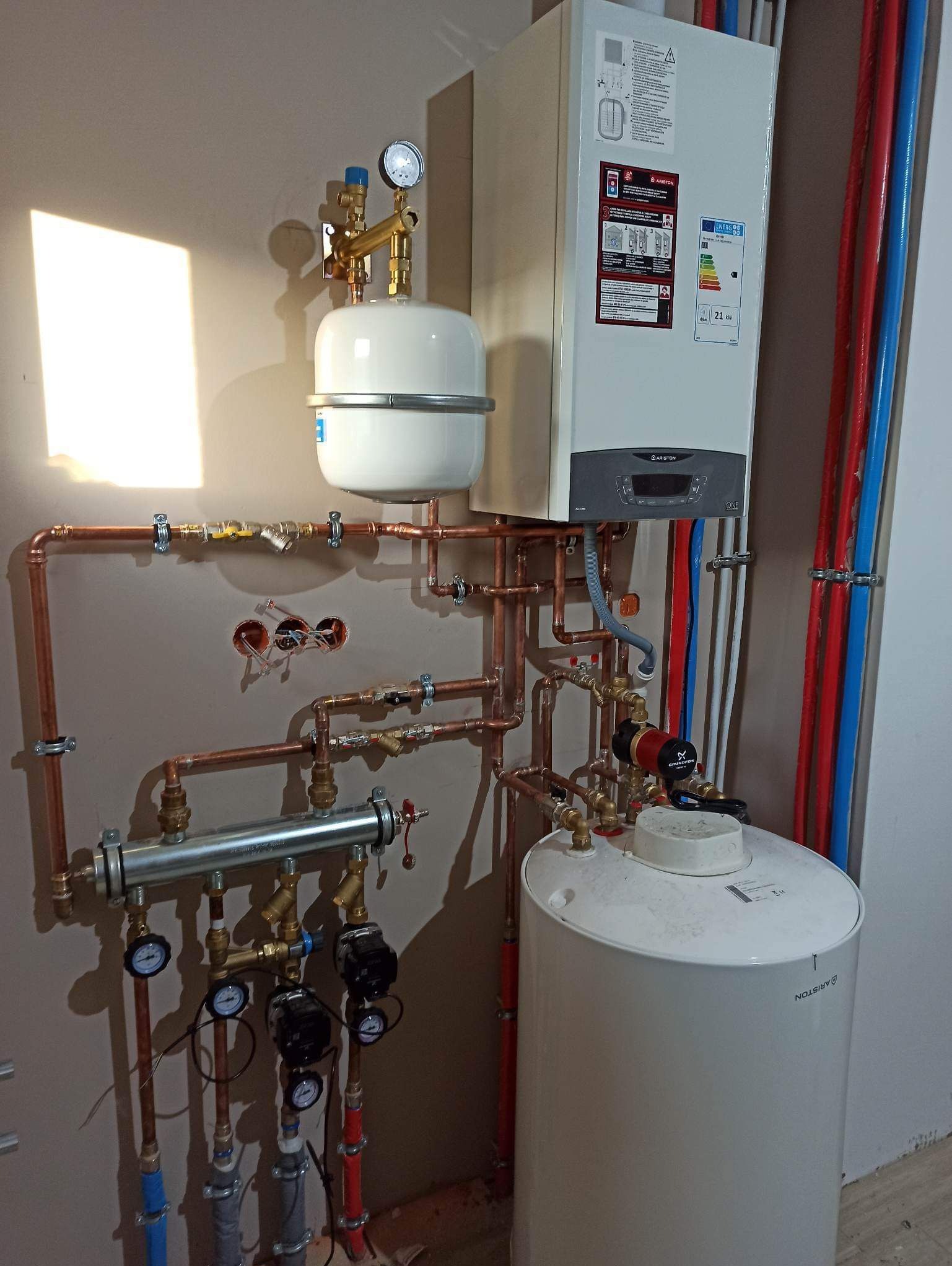 Nowoczesny piec gazowy Ariston z kompletną instalacją hydrauliczną, obejmującą zbiornik wyrównawczy, pompę obiegową Grundfos, liczne zawory i manometry, rury miedziane oraz kolorowe rury z tworzywa...