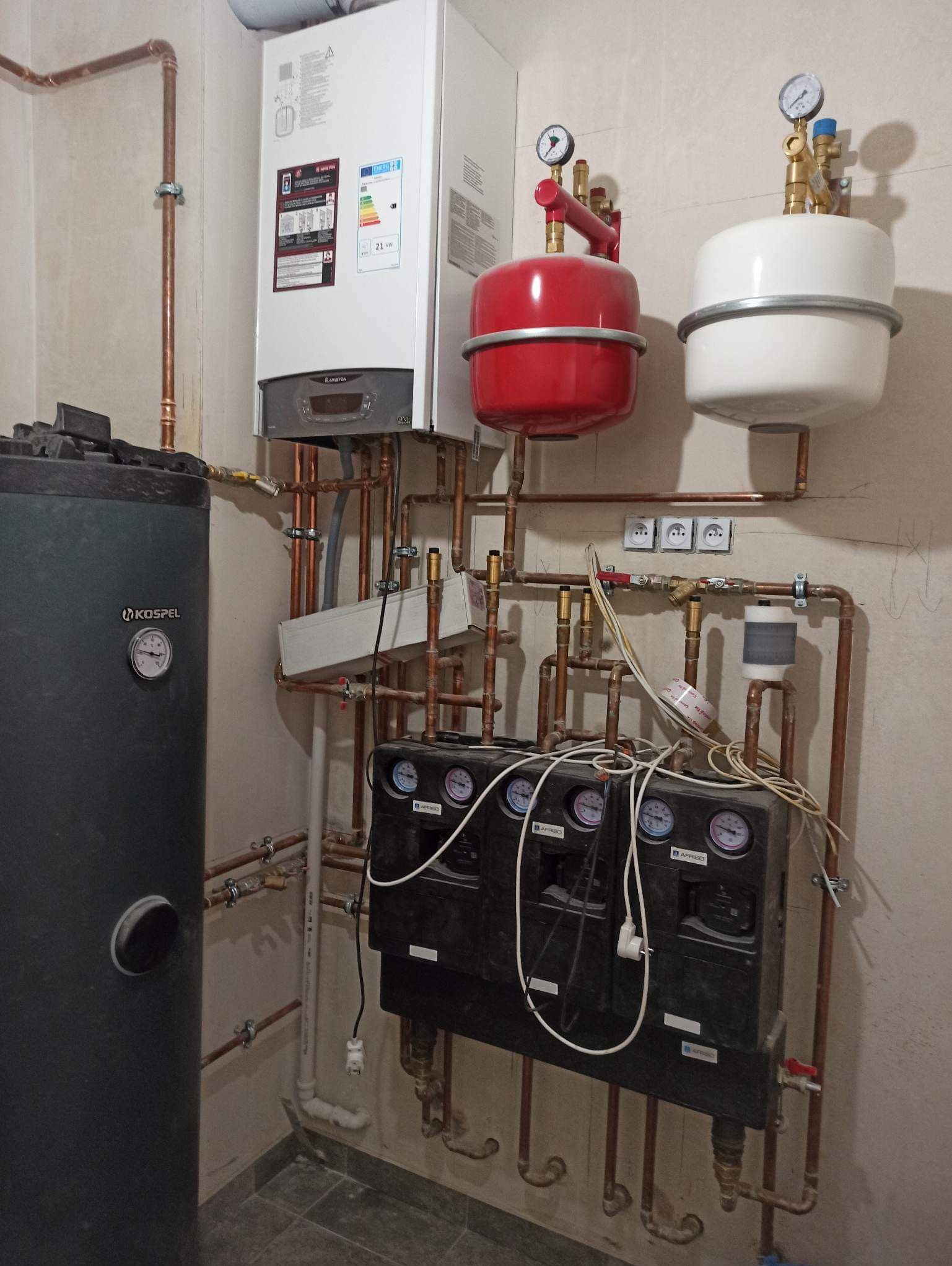 Złożona instalacja grzewcza z kotłem gazowym Ariston, czarnym zasobnikiem Kospel, czerwonym i białym naczyniem przeponowym oraz rozdzielaczem z manometrami Affriso, zamontowana na beżowej ścianie...