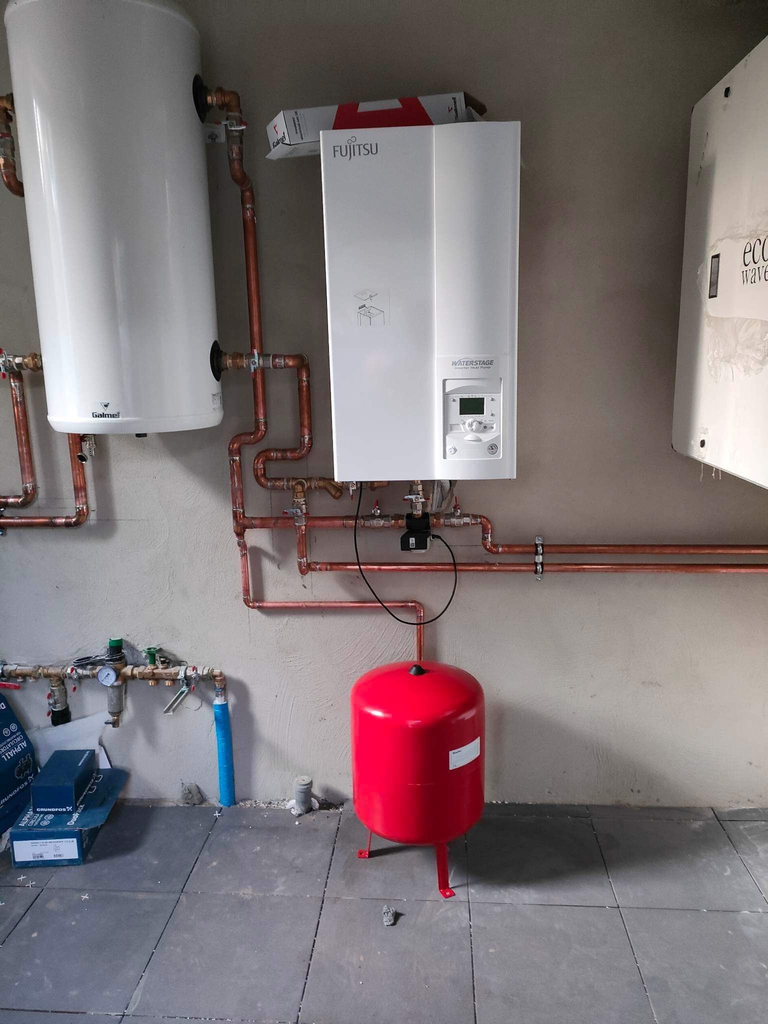 Instalacja CO z piecem gazowym Fujitsu Waterstage, zasobnikiem Galmet i czerwonym zbiornikiem wyrównawczym, rury miedziane prowadzone wzdłuż szarej ściany, widoczne pudełka Grundfos.