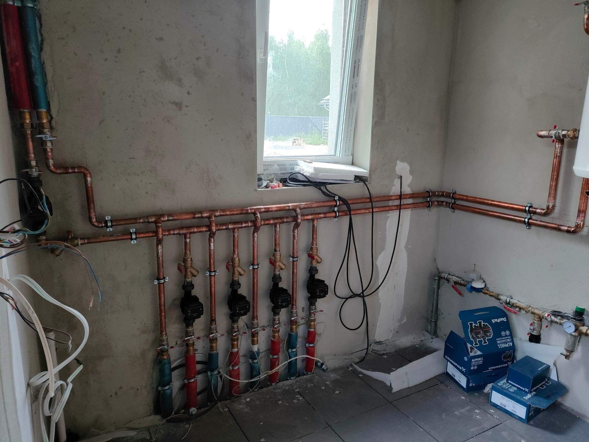Miedziane rury instalacji grzewczej z zaworami i pompami obiegowymi, częściowo zaizolowane, zamontowane na surowej ścianie w pomieszczeniu z oknem, widoczne opakowania pomp Grundfos.