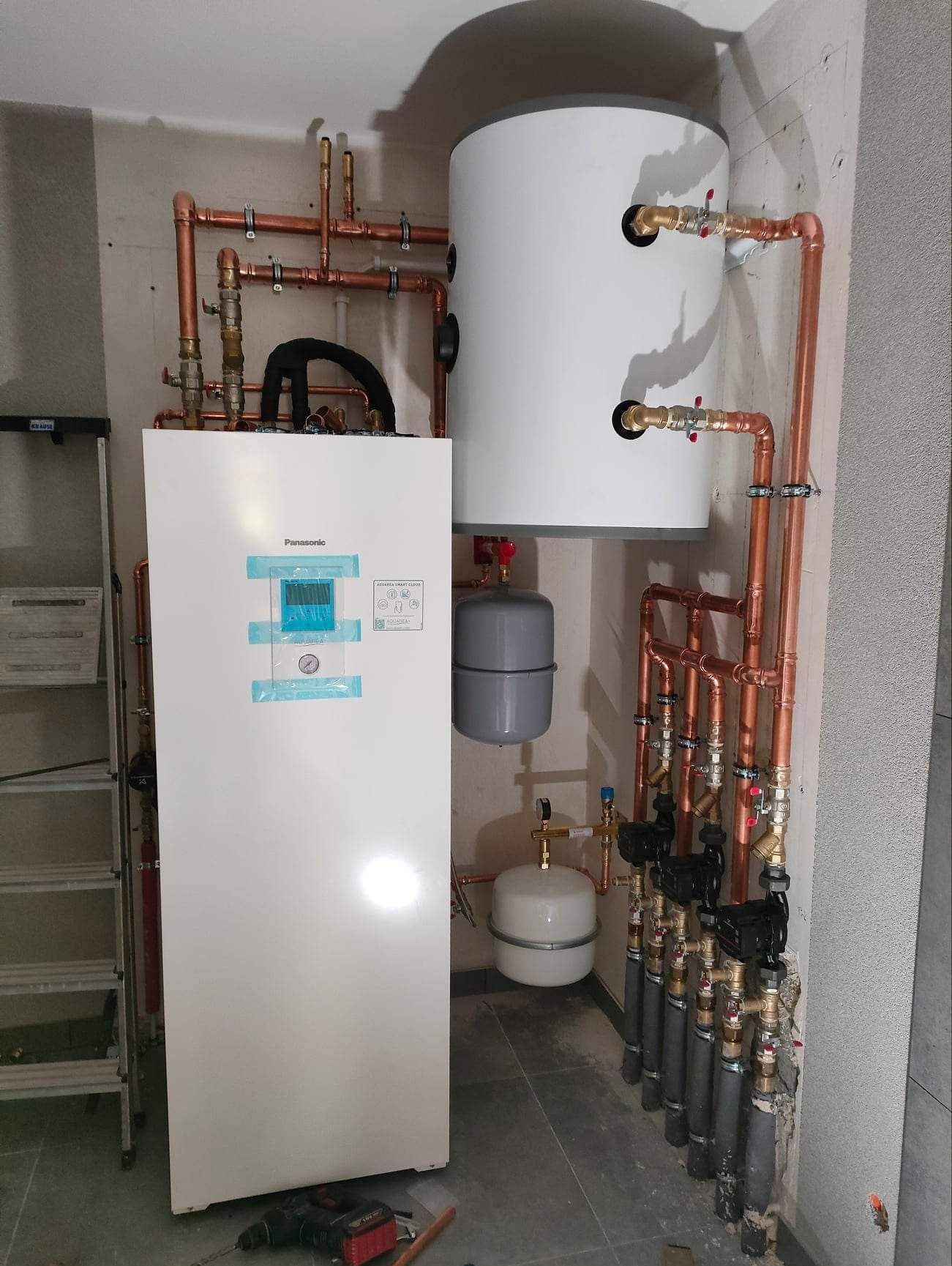 Instalacja pompy ciepła Panasonic Aquarea Smart Cloud z miedzianymi rurami, zbiornikiem buforowym i grupą pompową, widoczne narzędzia na podłodze.
