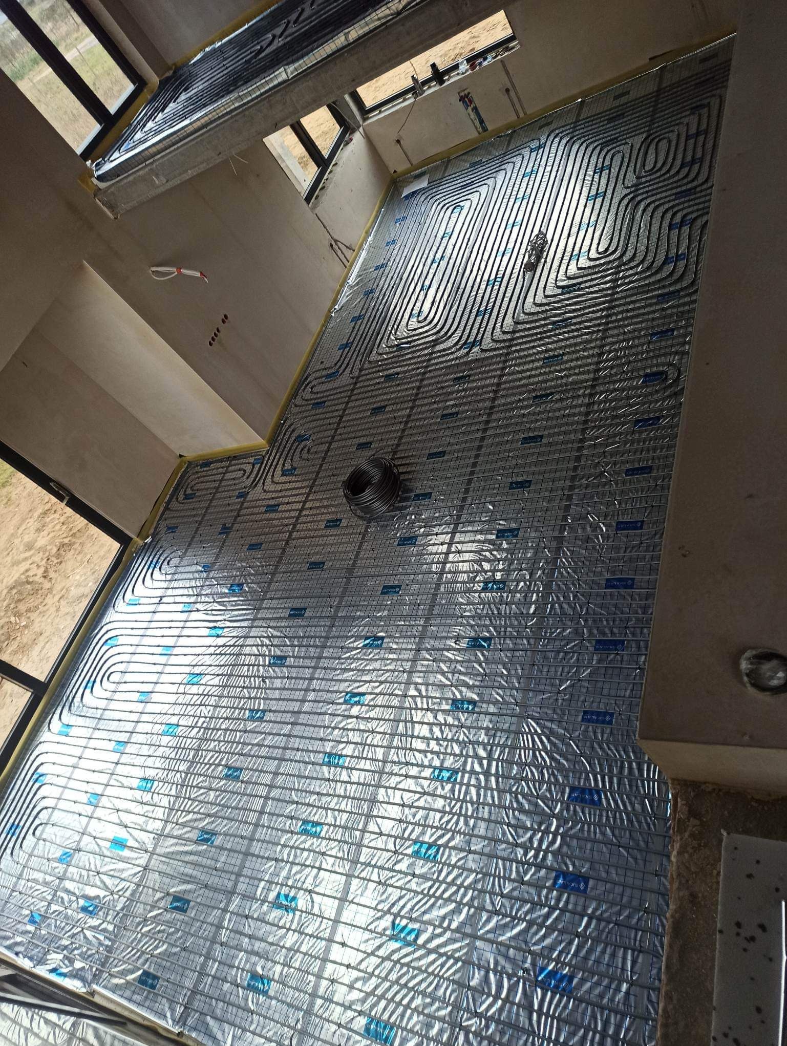 Rozłożona instalacja ogrzewania podłogowego na folii izolacyjnej z siatką, widok z góry na surowy stan wykończenia wnętrza z oknami.