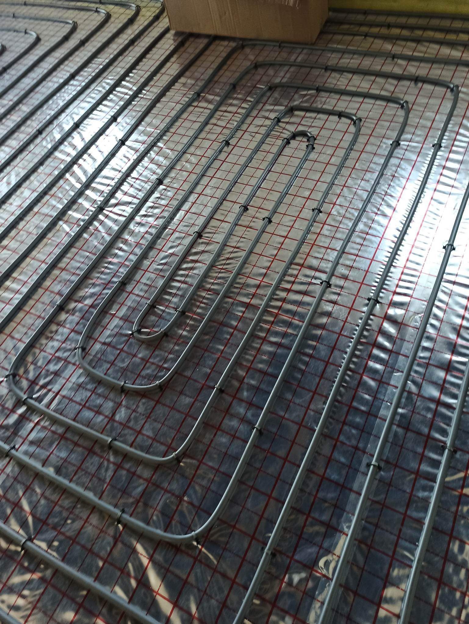 Szare rury ogrzewania podłogowego ułożone na folii z czerwoną siatką, zabezpieczone czarnymi klipsami, widok z góry.