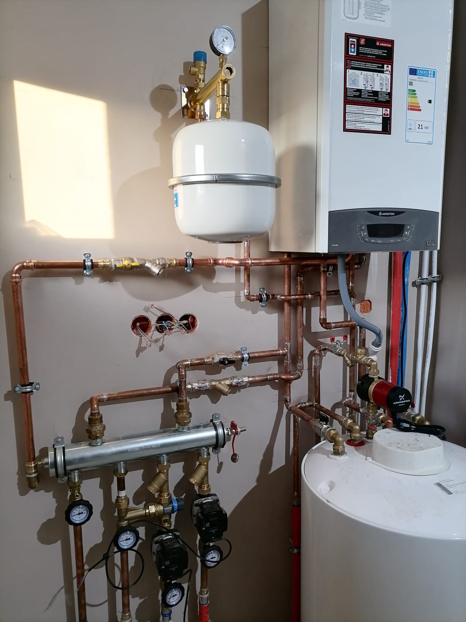 Nowoczesny piec gazowy Ariston ONE z rozbudowaną instalacją hydrauliczną z miedzianych rur, zbiornikiem wyrównawczym i pompą obiegową Grundfos, zamontowany na ścianie w jasnym pomieszczeniu.