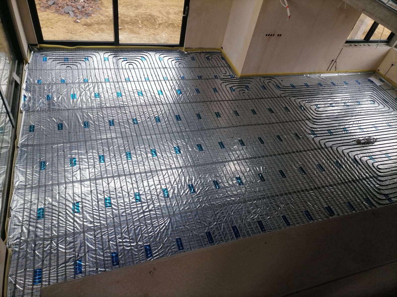 Instalacja ogrzewania podłogowego: rury grzewcze ułożone na folii aluminiowej z siatką, widok z góry, przygotowanie do zalania wylewką.