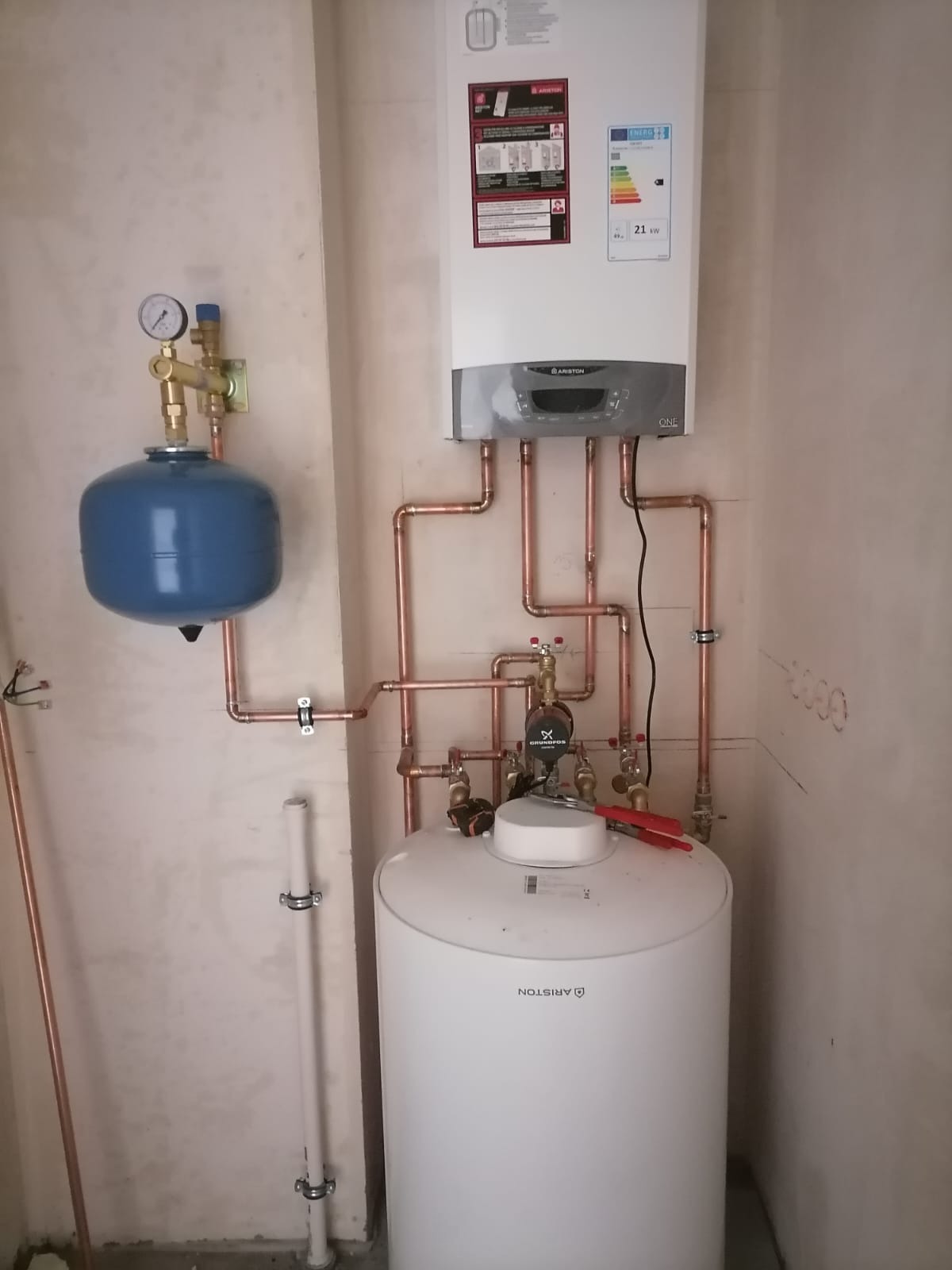 Nowoczesny piec gazowy Ariston z zasobnikiem ciepłej wody i niebieskim naczyniem przeponowym, zamontowany na ścianie z widocznymi miedzianymi rurami instalacji grzewczej.