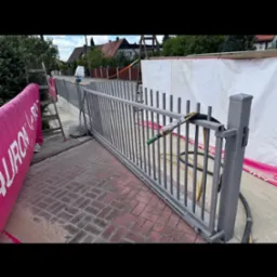 Szara metalowa brama po piaskowaniu, z widoczną różnicą w czystości kostki brukowej. W tle domy i zieleń. Dysza do piaskowania umieszczona na bramie.