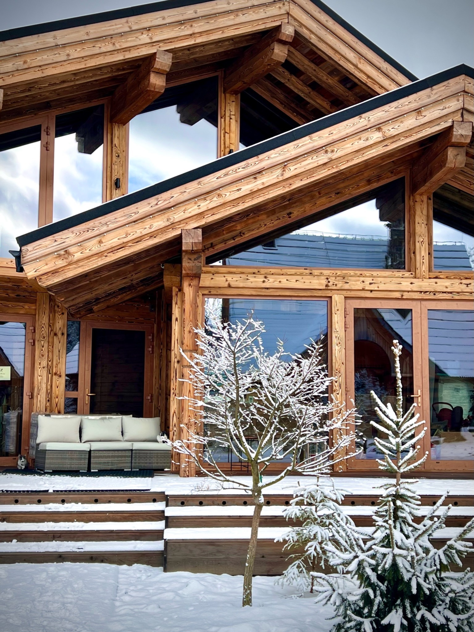 Drewniany dom z dużymi oknami i tarasem, zasypany śniegiem. Na tarasie sofa. Widoczne drzewka iglaste i liściaste pod śniegiem. Charakterystyczna konstrukcja dachu.