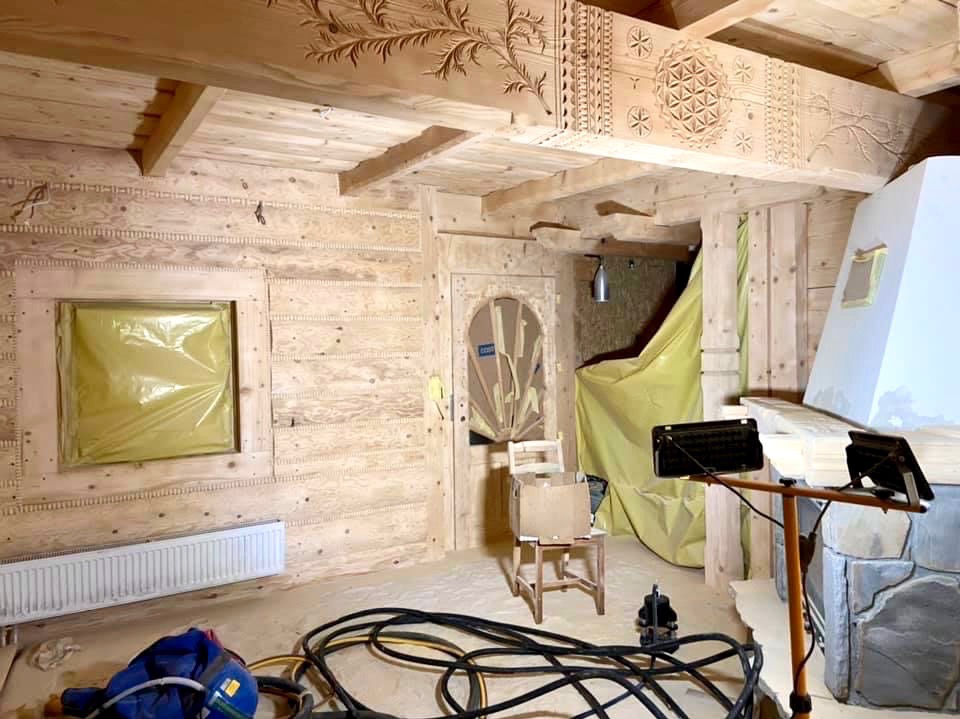 Wnętrze drewnianego domu w trakcie renowacji, z zakrytymi oknami, widocznym kominkiem i detalami zdobniczymi na belkach stropowych. Prace wykończeniowe w toku.