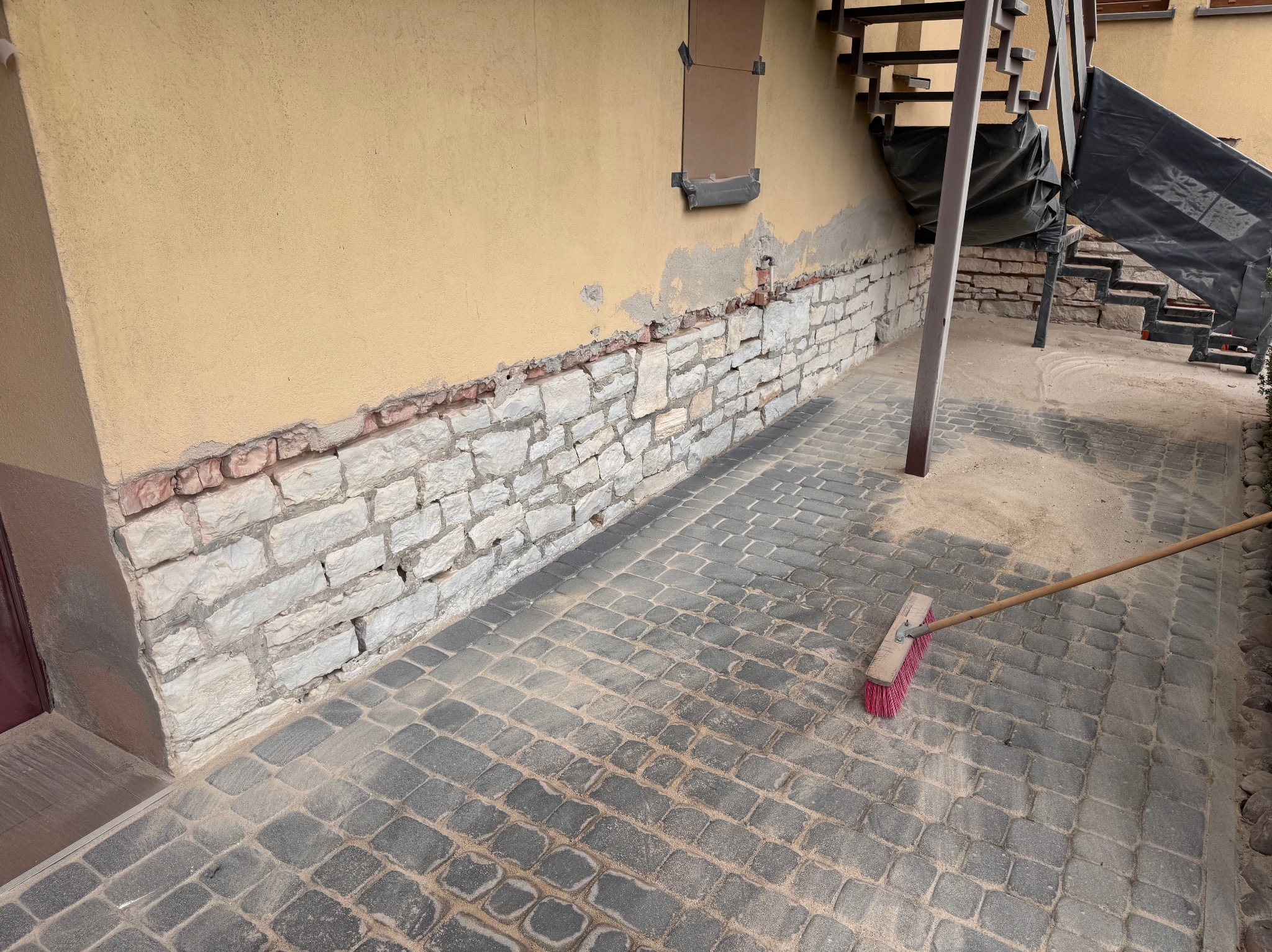 Oczyszczona kamienna elewacja budynku z fragmentami starego tynku i chodnik z kostki brukowej po piaskowaniu, zamiatanie resztek piasku, widoczne schody.