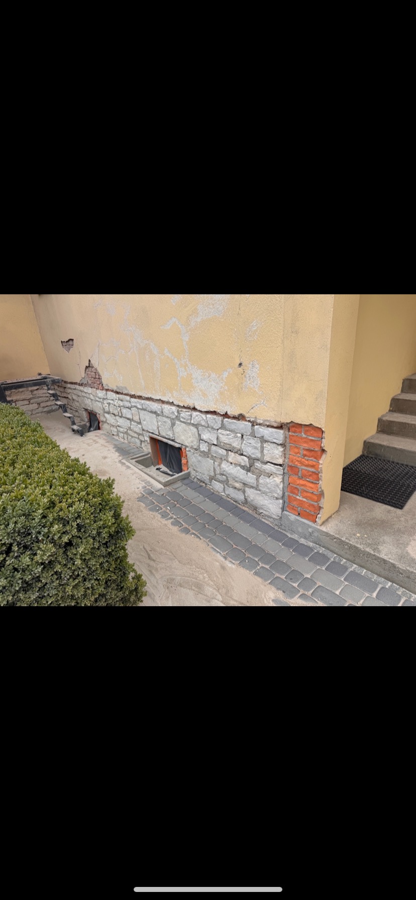 Elewacja budynku z fragmentami kamienia i cegły, widoczne ślady uszkodzeń i napraw. Częściowo odnowiona ściana, zieleń w tle, kostka brukowa.
