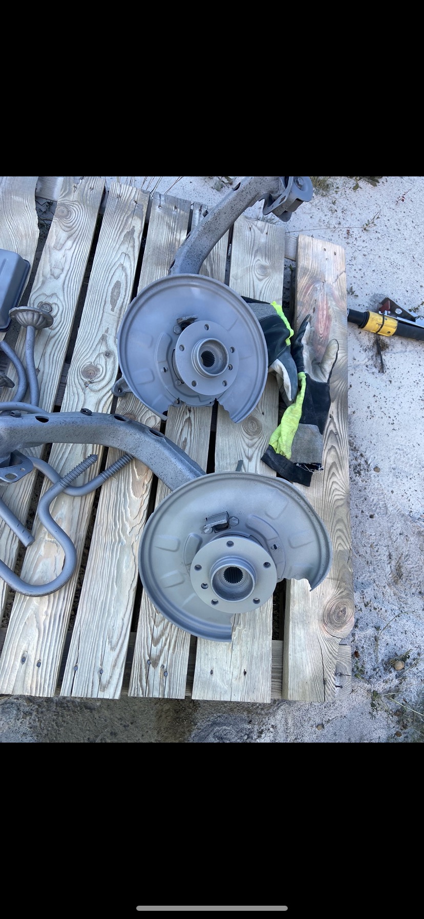 Metalowe elementy zawieszenia po piaskowaniu/malowaniu na drewnianej palecie, obok leżą rękawice robocze i pistolet do piaskowania.