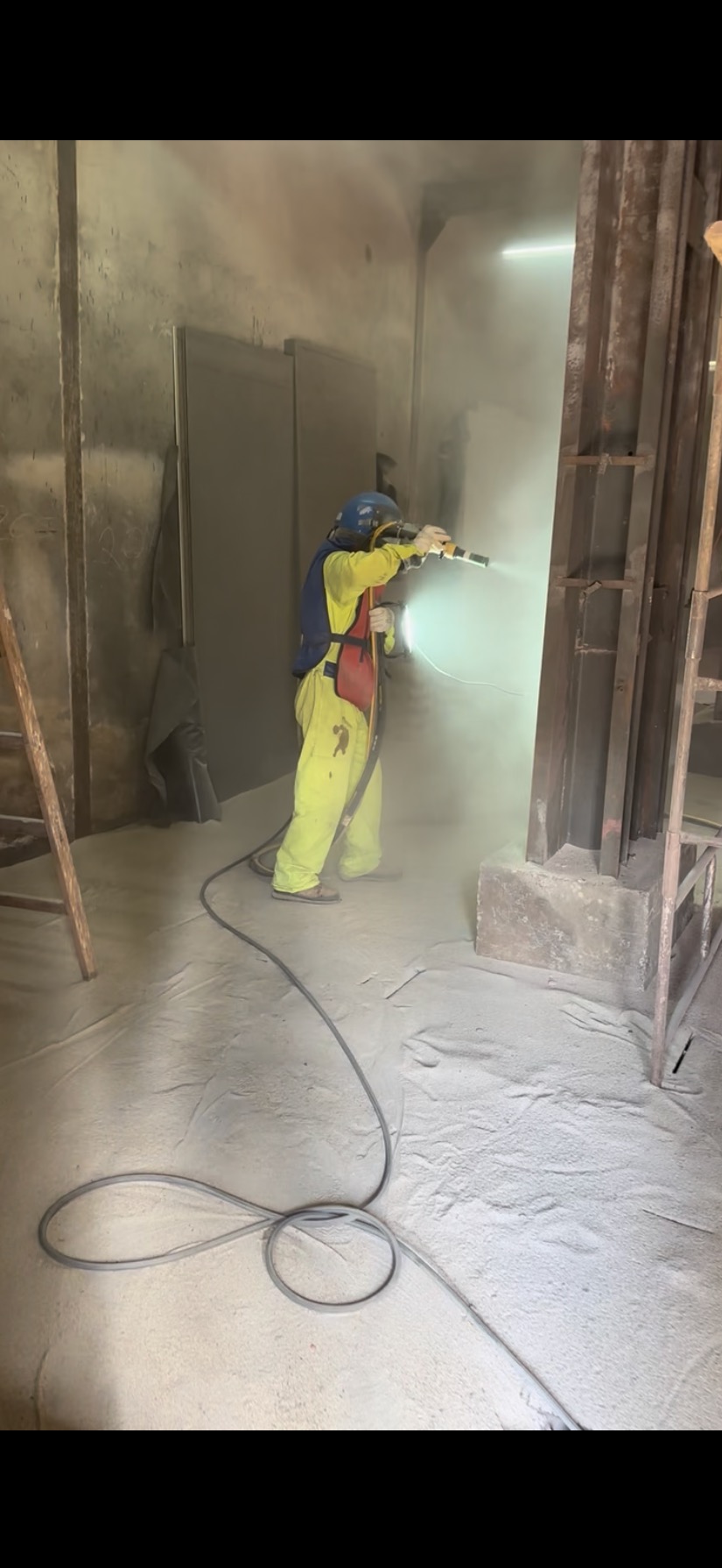 Pracownik w kombinezonie ochronnym piaskuje metalową konstrukcję wewnątrz hali. Widoczny pył i przewód. Oświetlenie górne.