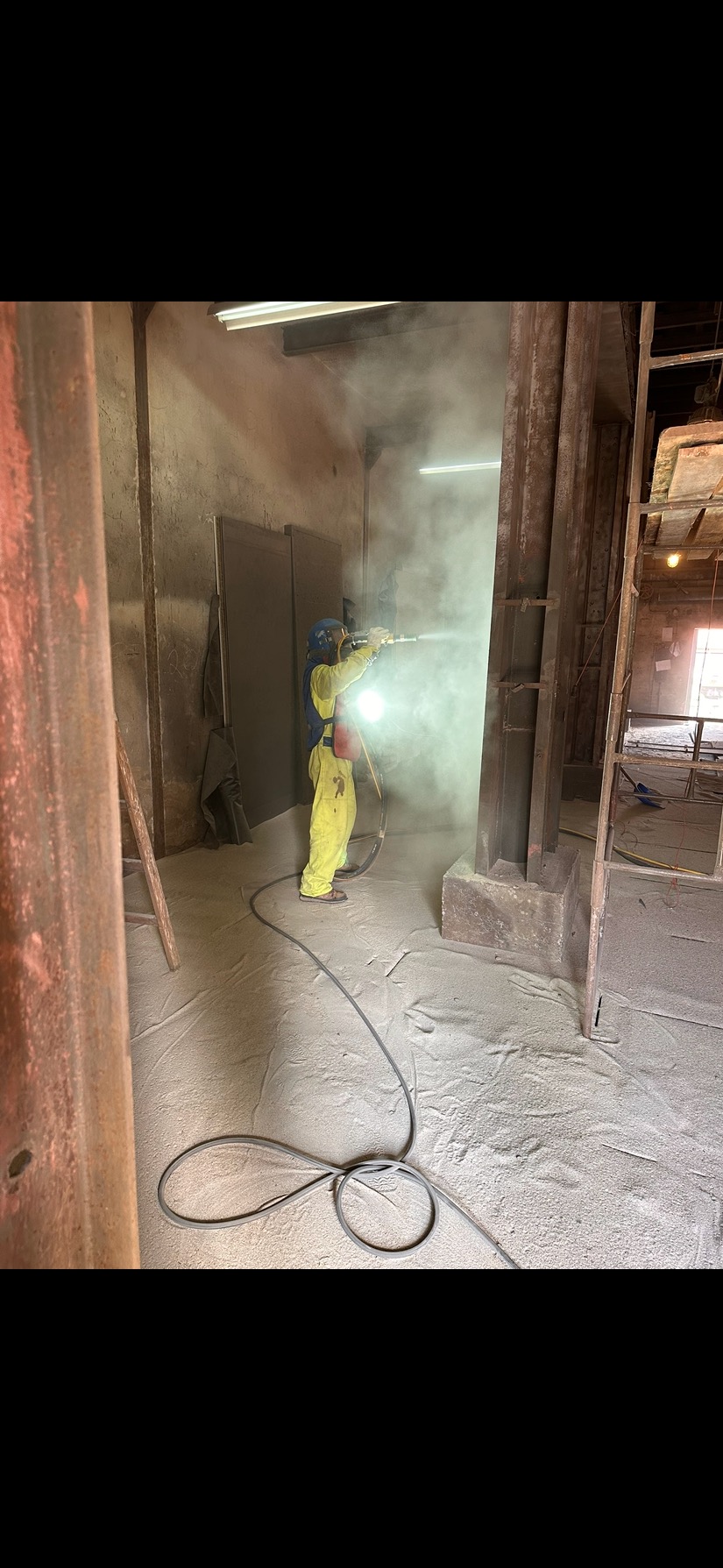 Pracownik w kombinezonie ochronnym piaskuje metalową konstrukcję wewnątrz budynku, unosząc tumany pyłu. Na podłodze warstwa piasku.