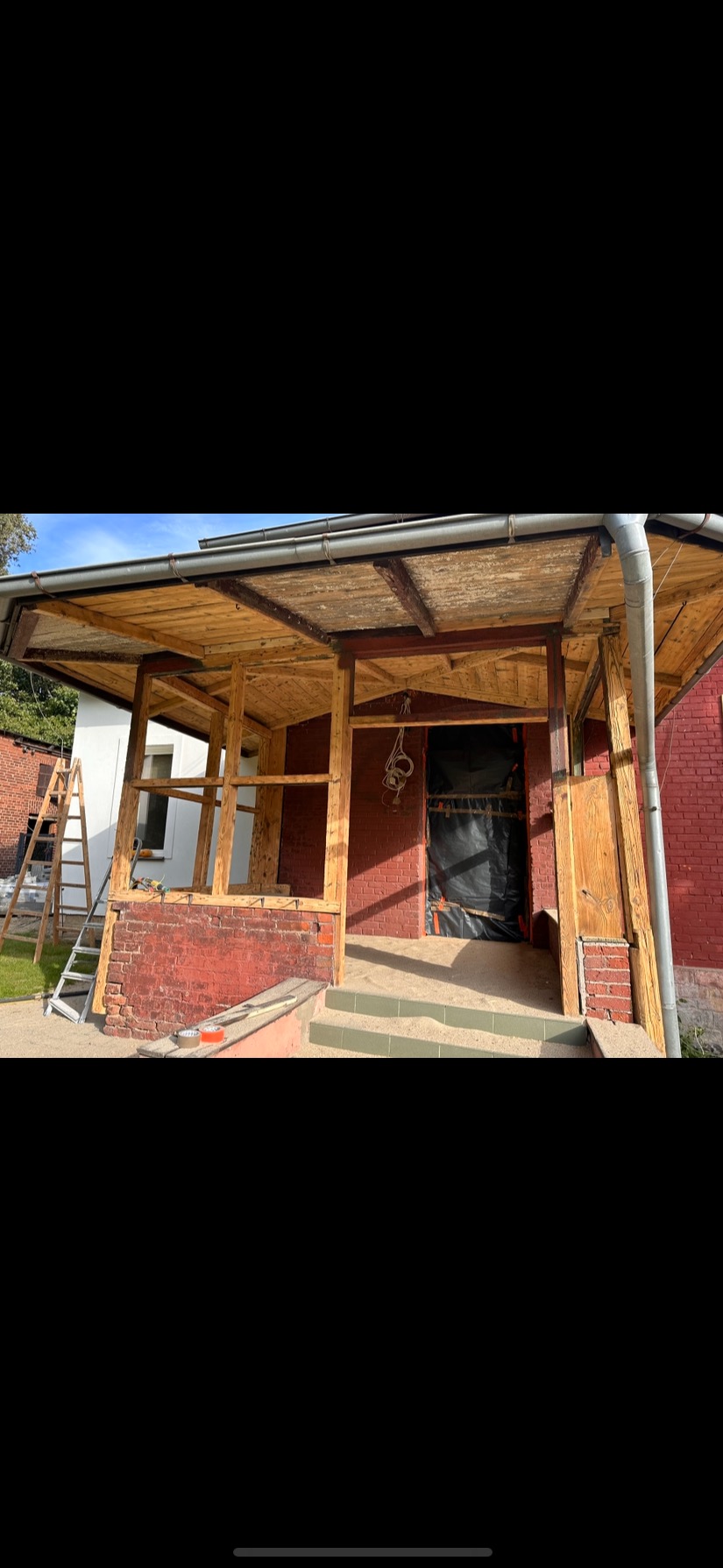 Remont werandy domu z czerwonej cegły. Widoczne drewniane elementy konstrukcyjne, drabina i narzędzia. Częściowo zakryte wejście folią ochronną. Etap prac w toku.