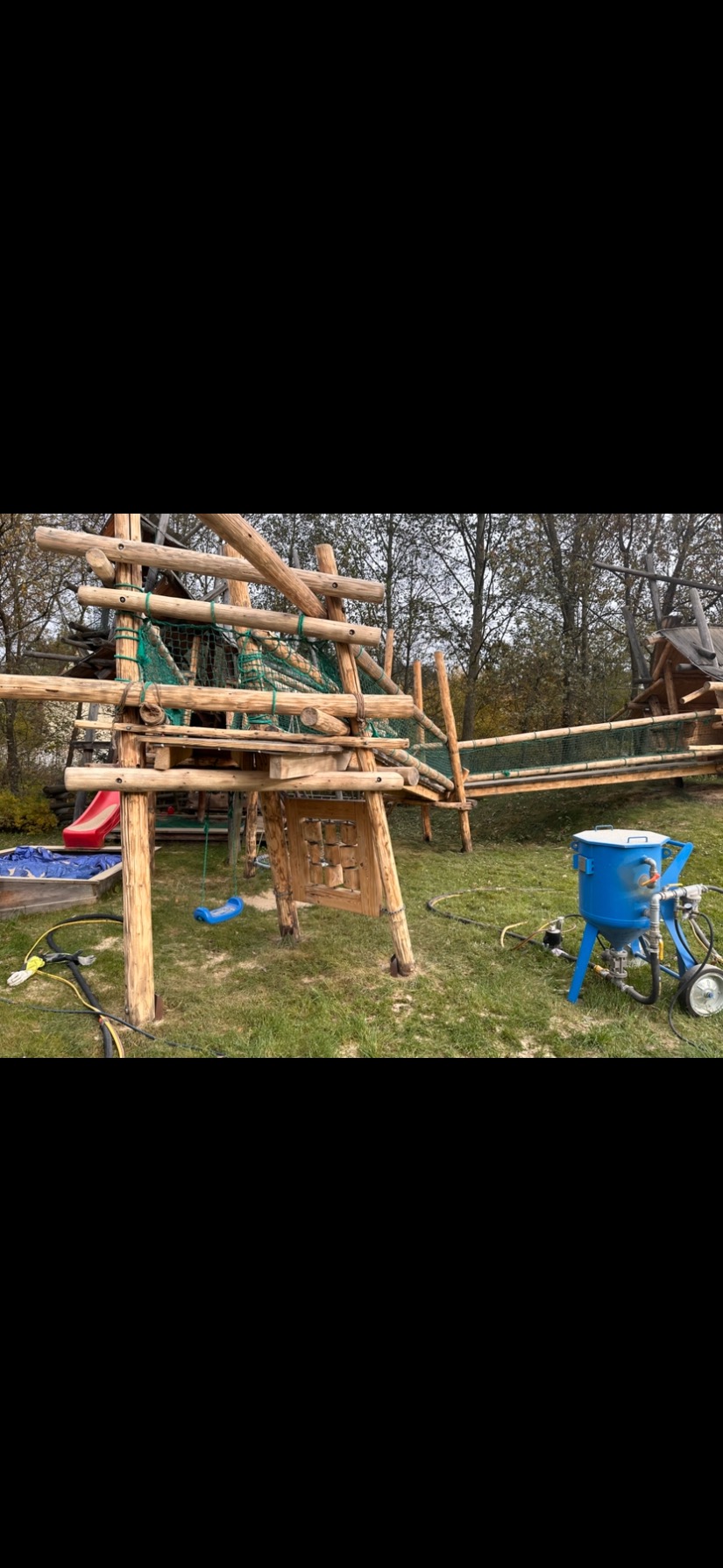 Piaskowanie drewnianego placu zabaw na zewnątrz. Widoczna piaskarka, elementy konstrukcji z surowego drewna, siatka, zjeżdżalnia i huśtawka.