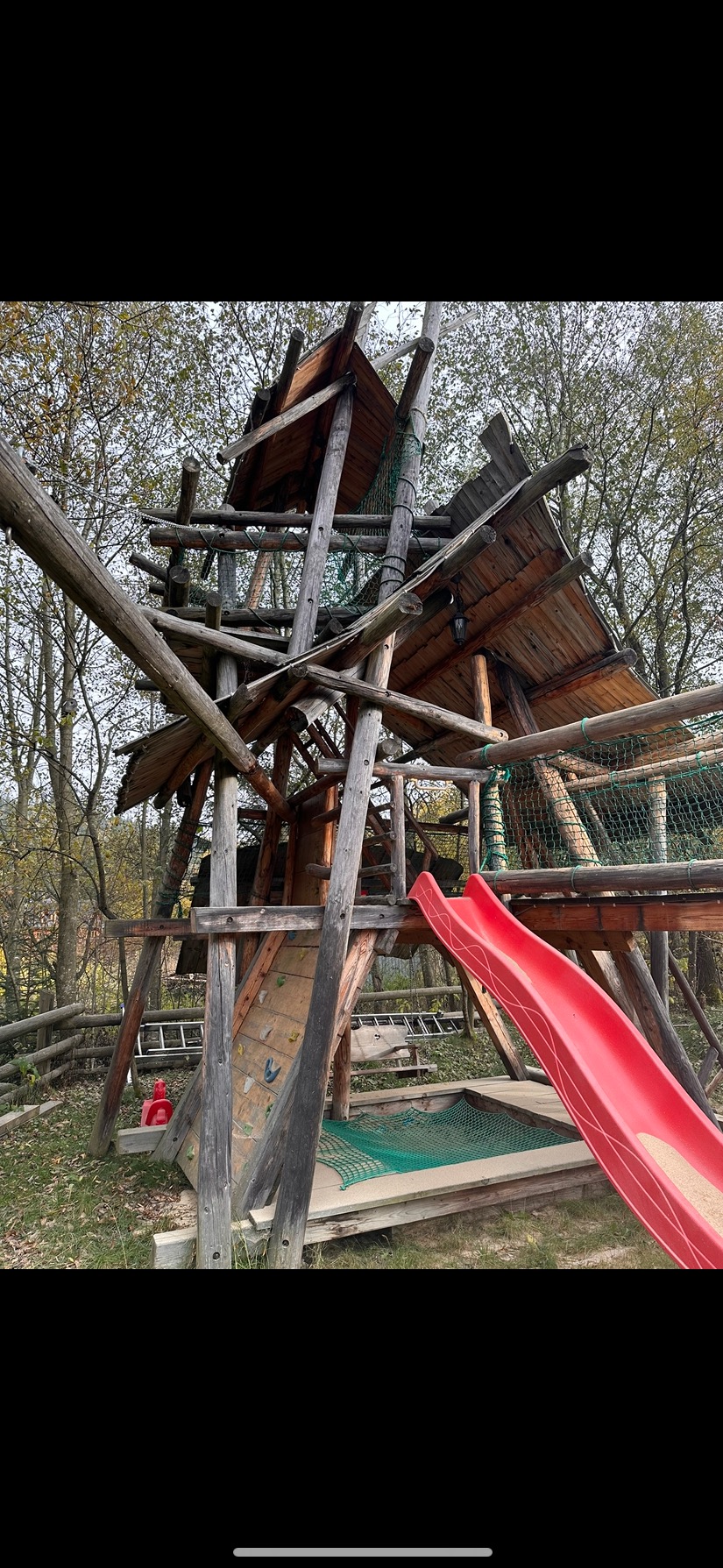 Drewniany plac zabaw z czerwoną zjeżdżalnią, siatkami i ścianką wspinaczkową na tle drzew. Konstrukcja z bali i desek, w naturalnym otoczeniu.