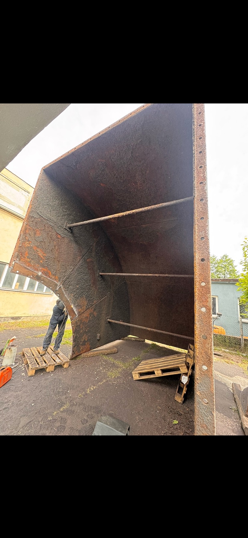 Piaskowanie wielkogabarytowego elementu konstrukcyjnego przez pracownika w kombinezonie, na zewnątrz. Widoczne ślady rdzy i palety.