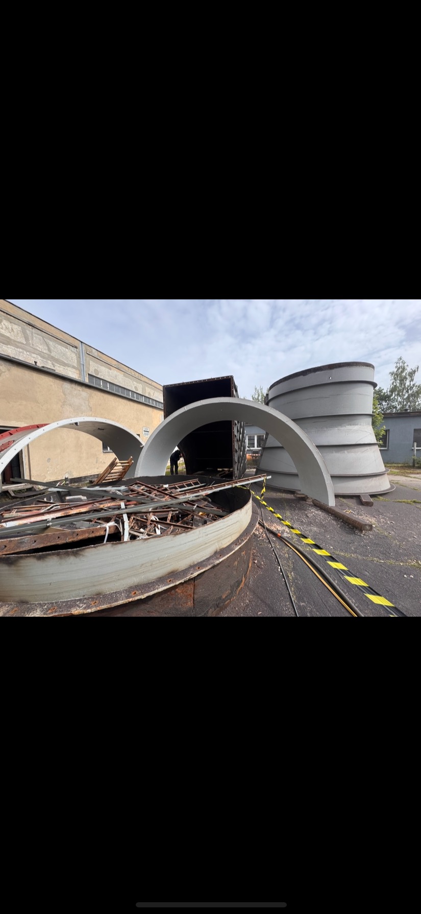 Elementy konstrukcyjne, prawdopodobnie po piaskowaniu, leżące na zewnątrz. Widoczny fragment budynku i kontener. Żółto-czarna taśma ostrzegawcza.