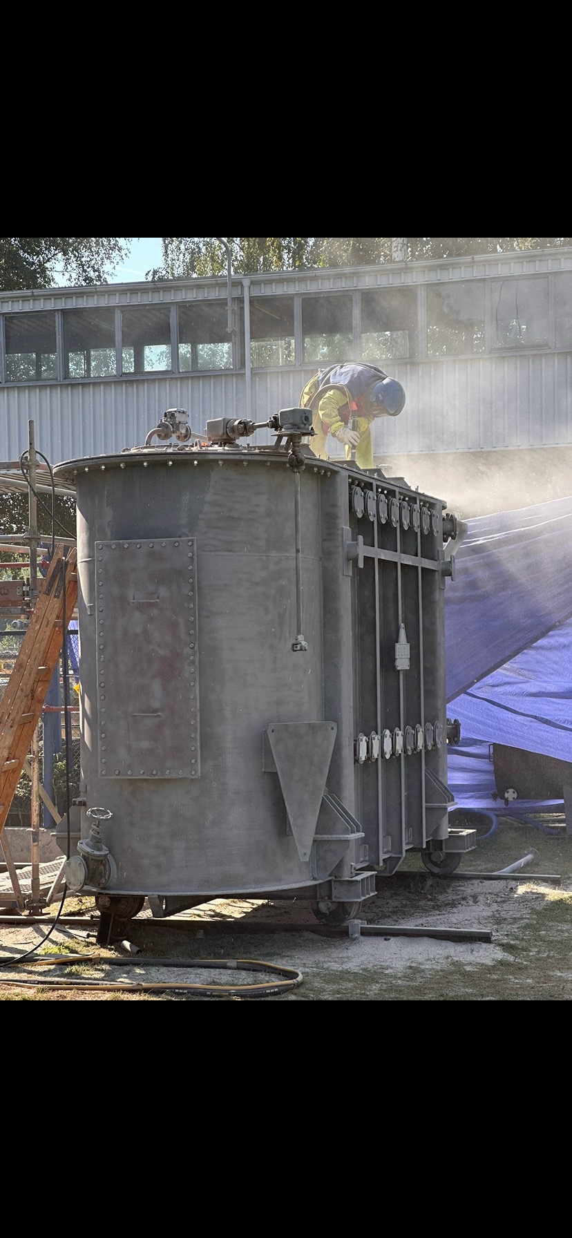 Pracownik w kombinezonie piaskuje duży, metalowy zbiornik przemysłowy na zewnątrz, z użyciem specjalistycznego sprzętu. W tle budynek i drabina.