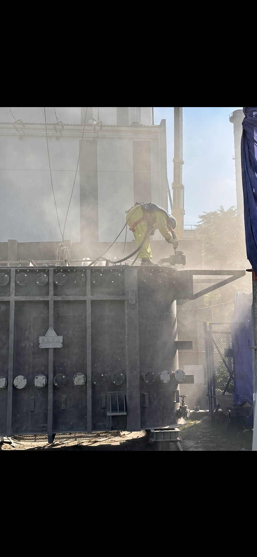 Pracownik w kombinezonie ochronnym piaskuje duży, metalowy transformator na zewnątrz, z widocznym pyleniem i elementami konstrukcji w tle.