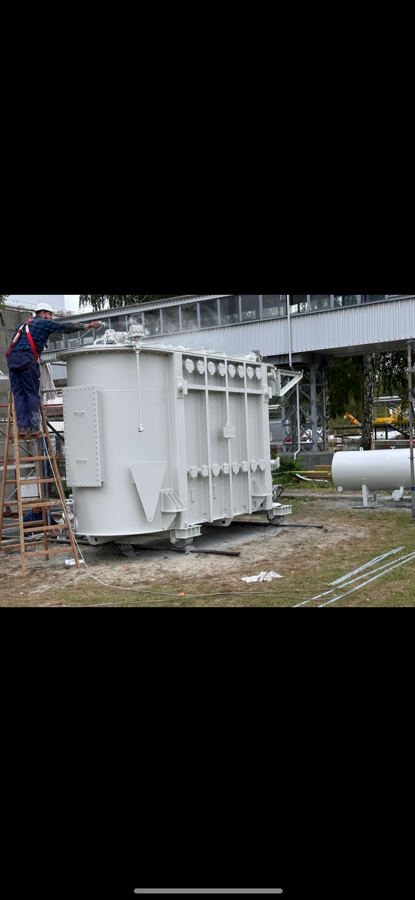 Pracownik w kasku maluje na biało duży transformator na zewnątrz, stojąc na drabinie. Widoczny most pieszy w tle i biały zbiornik.