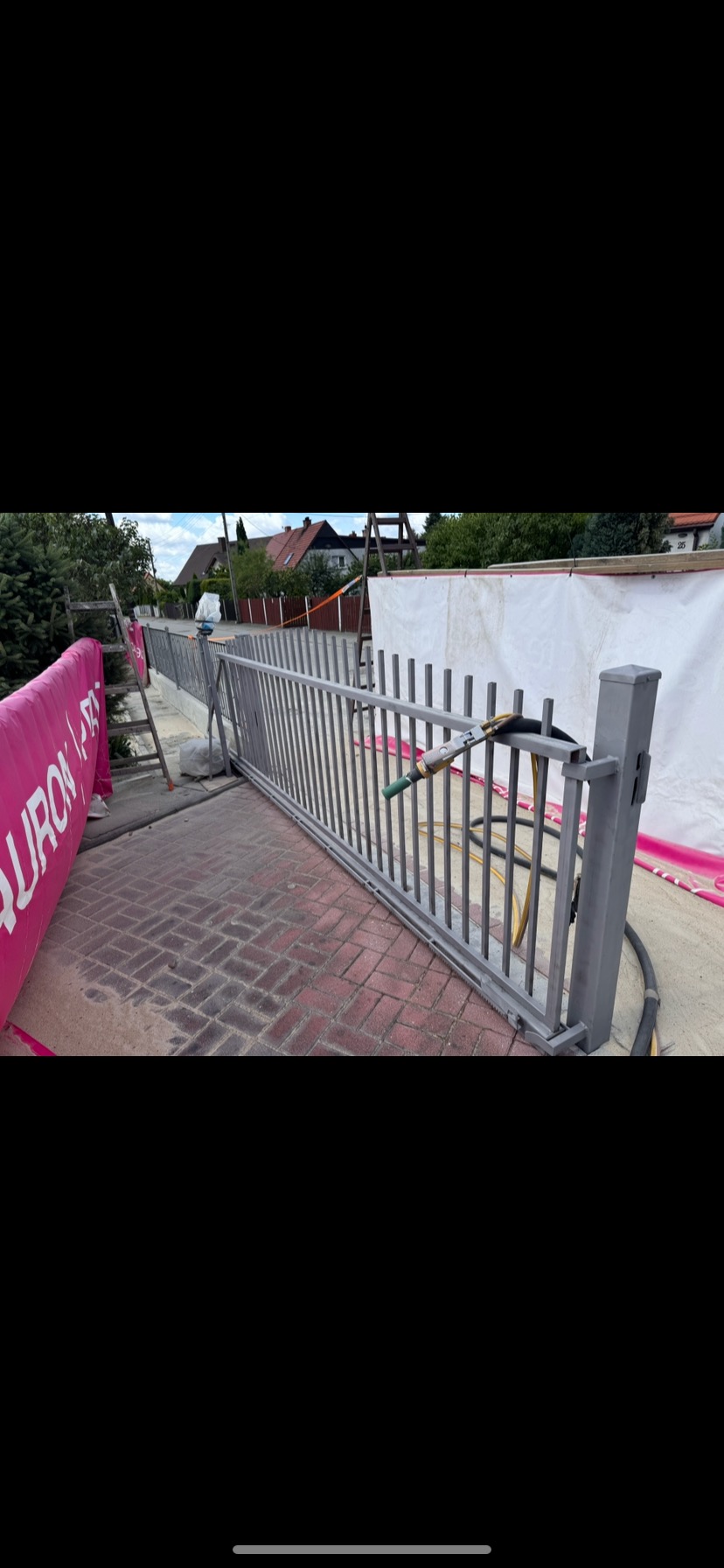 Szara metalowa brama po piaskowaniu, z widoczną różnicą w czystości kostki brukowej. W tle domy i zieleń. Dysza do piaskowania umieszczona na bramie.