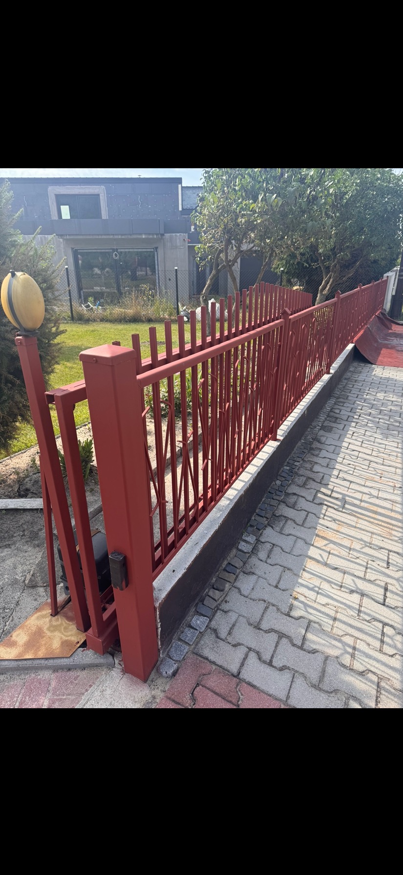 Czerwone, metalowe ogrodzenie posesji z bramą automatyczną, widoczne z poziomu chodnika. W tle budynek mieszkalny i zieleń.