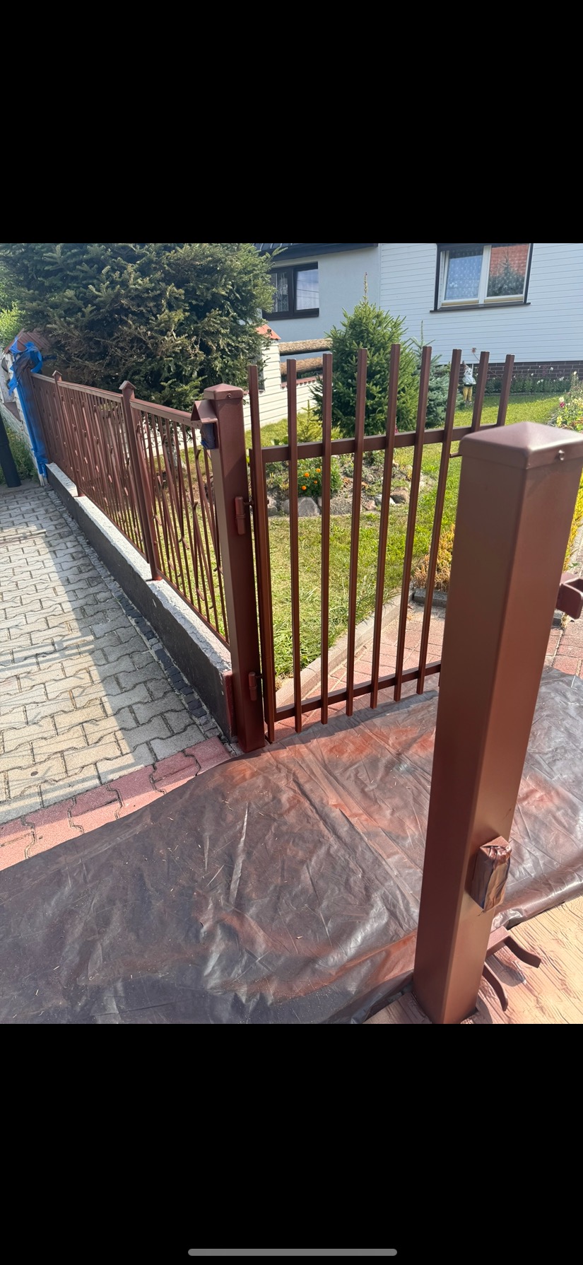 Świeżo pomalowane, brązowe ogrodzenie posesji z metalowymi sztachetami i słupkami na tle domu z ogrodem. Malowanie w toku, folia ochronna na podłożu.