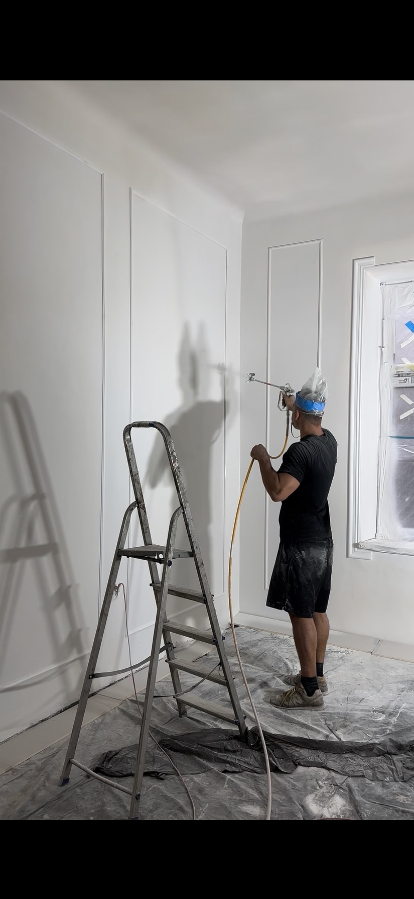 Malowanie ścian z dekoracyjnymi listwami natryskowo. Mężczyzna w masce maluje białą farbą wnętrze pomieszczenia. Obok stoi drabina malarska. Podłoga zabezpieczona folią.