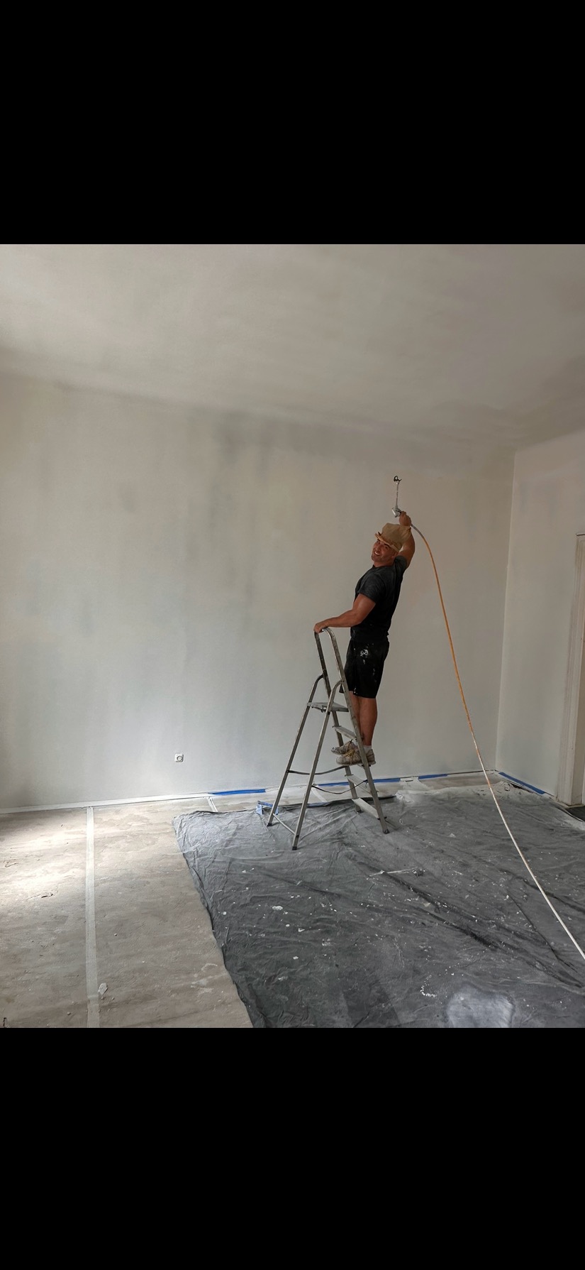 Malarz natryskowy na drabinie maluje ścianę w jasnym pokoju. Podłoga zabezpieczona folią malarską. Widoczne detale wykończeniowe, np. listwy przypodłogowe.