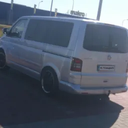 Srebrny bus Volkswagen Multivan zaparkowany na brukowanej powierzchni, widoczny z tyłu, z tablicą rejestracyjną 'R-Transport', na tle budynku Makro.