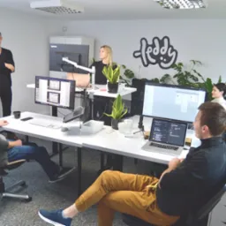 Zespół w biurze, omawiający projekt strony internetowej widoczny na ekranach komputerów, z logo firmy na ścianie i roślinami doniczkowymi.