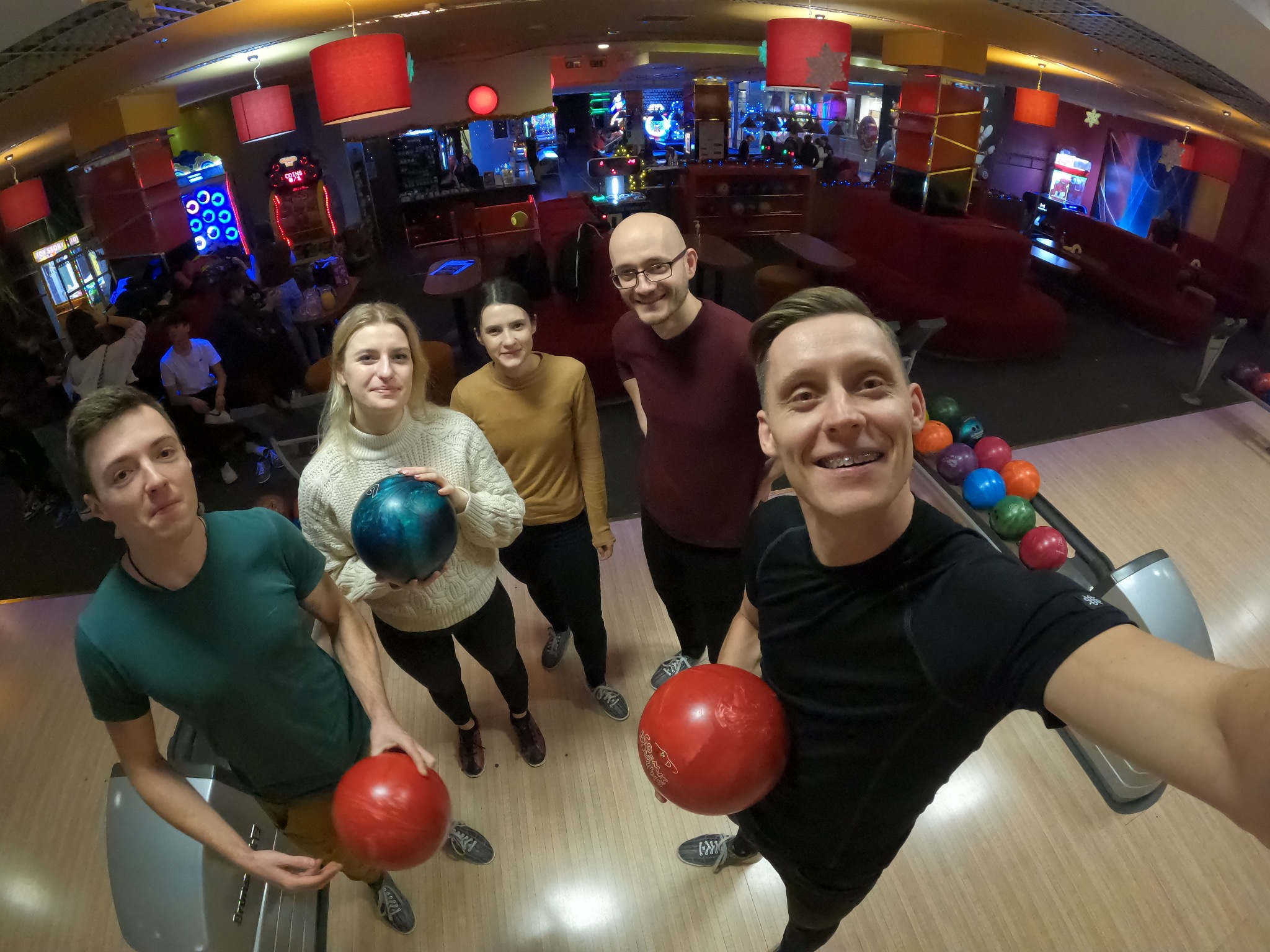 Grupa pięciu osób robiąca selfie w kręgielni, trzymają kule do kręgli, w tle automaty do gier.