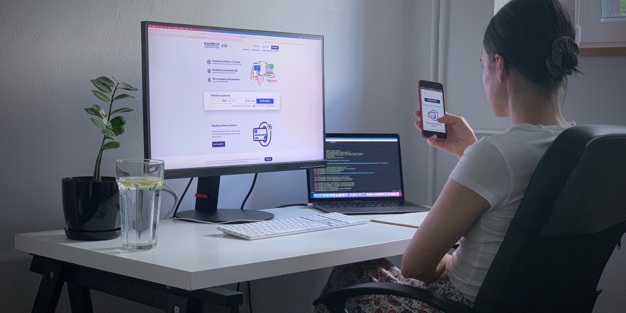 Kobieta przegląda stronę internetową transfer24 na telefonie, w tle monitor z otwartą stroną internetową i laptop z kodem źródłowym.