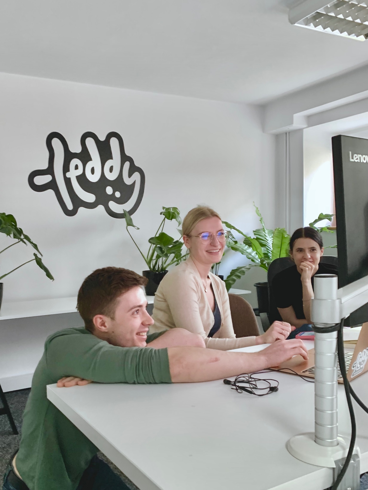 Zespół trzech osób w jasnym biurze, jeden mężczyzna opiera się o biurko, dwie kobiety siedzą przy komputerze, na ścianie logo firmy i rośliny doniczkowe.