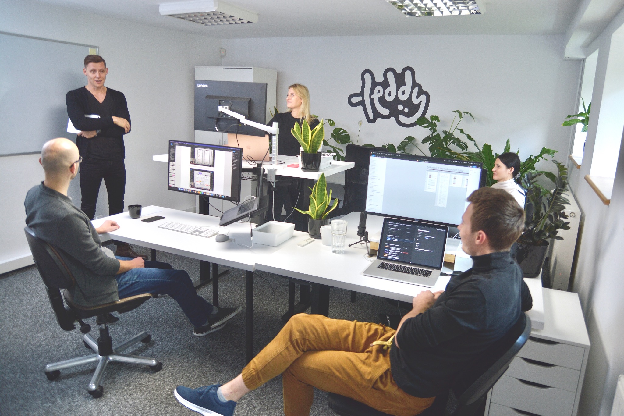 Zespół w biurze, omawiający projekt strony internetowej widoczny na ekranach komputerów, z logo firmy na ścianie i roślinami doniczkowymi.