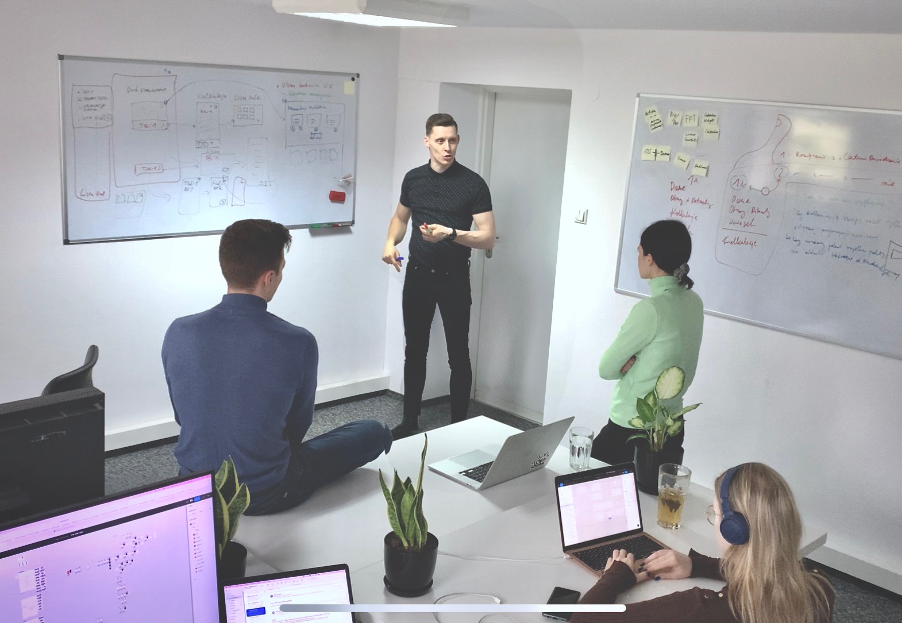Zespół podczas burzy mózgów w biurze, omawiający schematy i plany na tablicach, z laptopami i roślinami na stole, przy naturalnym oświetleniu.