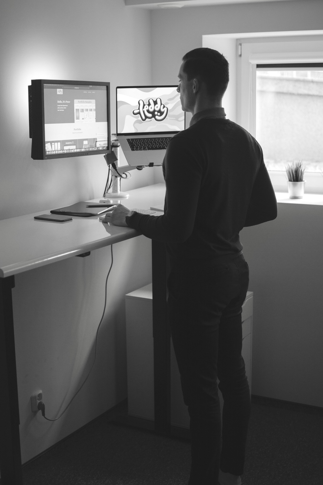 Mężczyzna stoi przy biurku z podnoszonym blatem, pracując na laptopie z wyświetlonym logo i monitorze z widoczną stroną internetową. Ustawienie w biurze, widok z tyłu, czarno-białe zdjęcie.