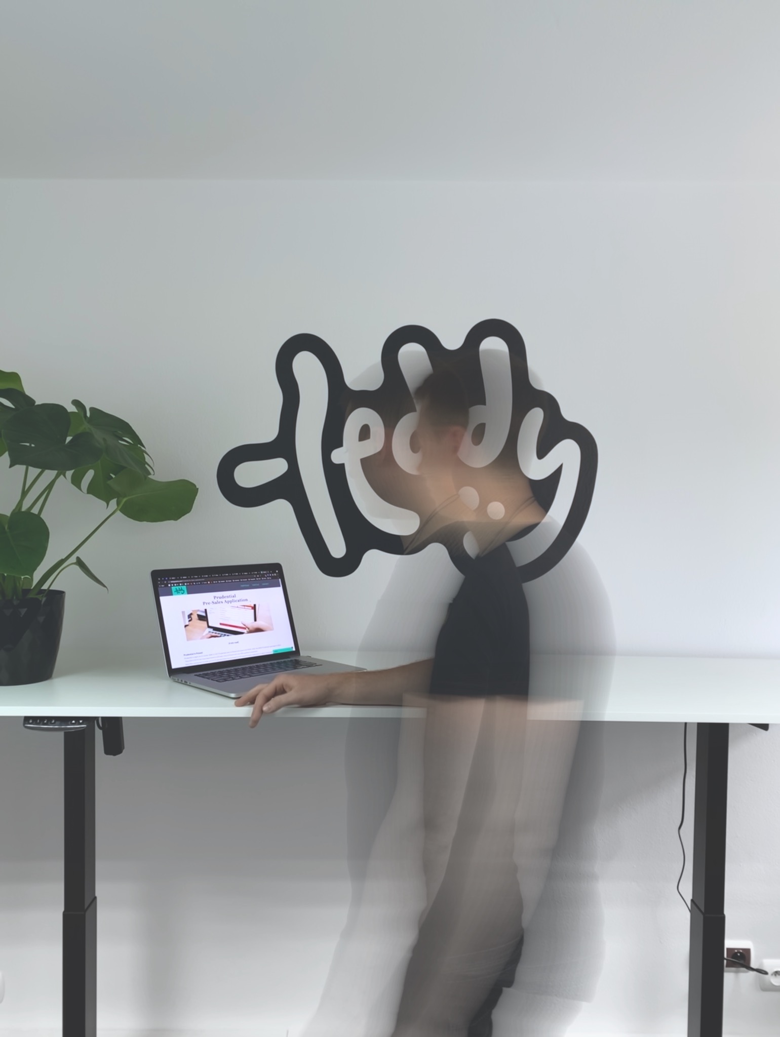 Osoba w ruchu siedzi przy regulowanym biurku z laptopem, na ekranie widoczna strona internetowa, za nią na ścianie dekoracyjny napis w stylu logo, obok stoi roślina doniczkowa.