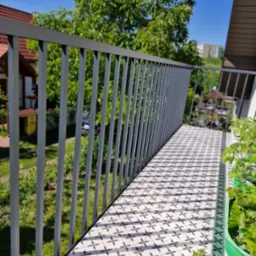 Nowoczesna, szara balustrada balkonowa z pionowych profili, wzorzysta podłoga balkonu w biało-czarne kwiaty, widok na zieleń ogrodu i zabudowania w tle, donice z sadzonkami z prawej strony.